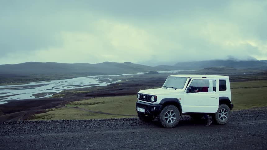 Woman with backpack gets into offroad vehicle on gravel road surrounded by hills and a winding river
