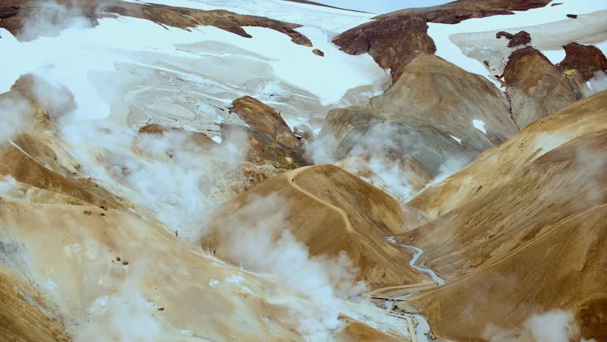 Steam vents rise from a rugged snow capped geothermal valley with colorful slopes and a winding stream