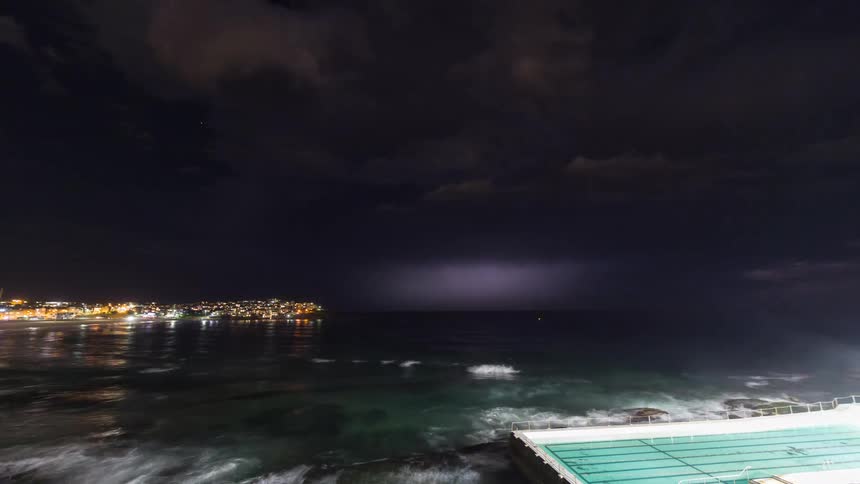 Lightning strikes above the ocean near a brightly lit city at night