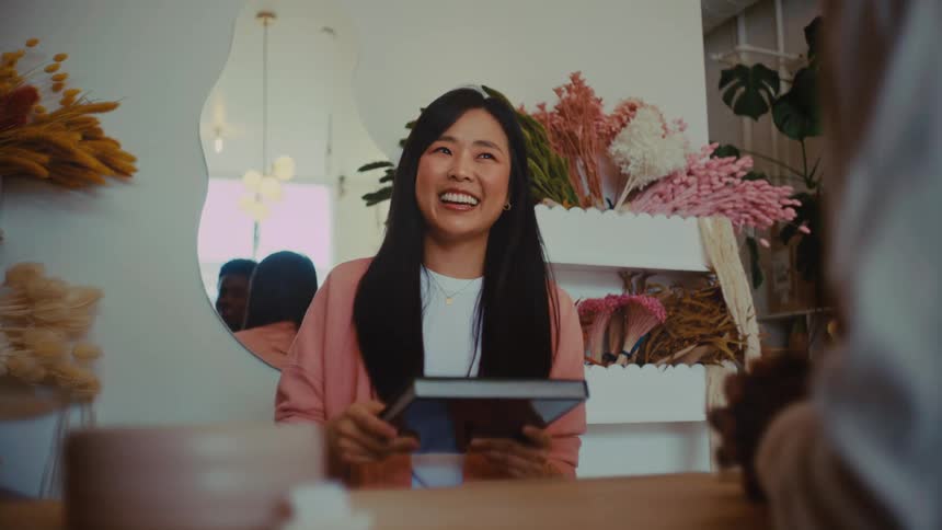 A cashier stands at the counter at midday handing a notebook to a customer with a warm smile they share a brief friendly exchange as the light from the storefront windows brightens the space around them