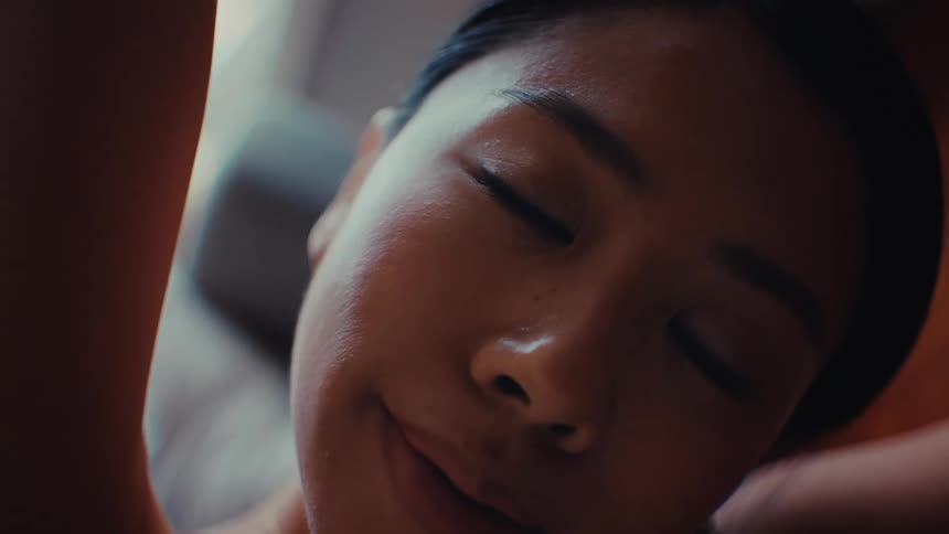 A close view of a young woman with her eyes closed as she sits in a room and smiles while tilting her head