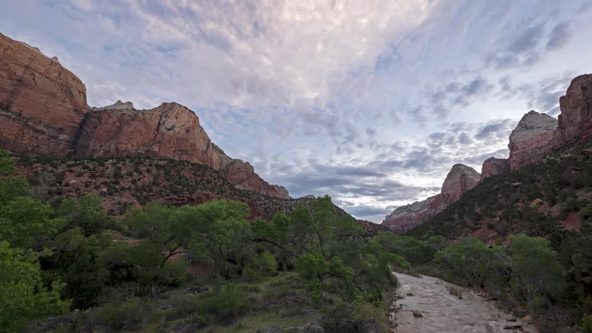 A stream flows through a rocky formation at midday clouds drifting slowly overhead a thin path of grass grows along the side of the water adding a soft line of green to the rugged scene