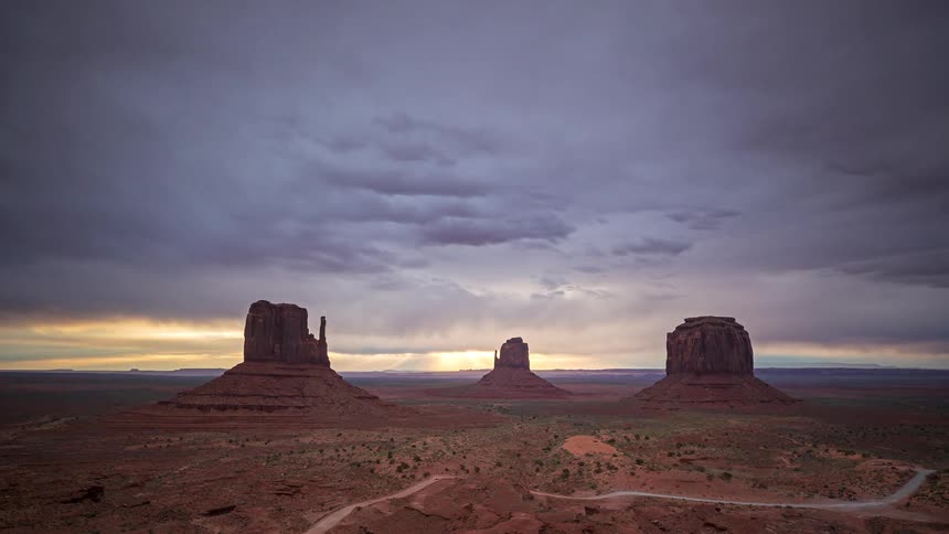 Cars travel on a road near buttes and rock formations in the desert as clouds move overhead