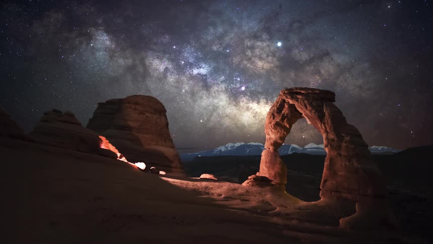 The milky way galaxy glows brightly in the night sky above a desert landscape while distant snow capped mountains sit on the horizon and artificial light illuminates the surrounding desert sand and rock