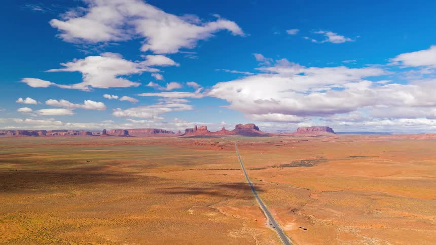 This high angle aerial shot shows a long straight highway cutting through a vast orange desert under a bright blue sky filled with drifting white clouds and soft shadows on the ground
