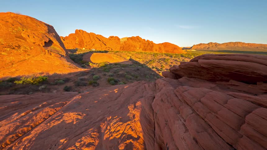 The camera moves over textured red sandstone during sunset revealing a vast desert valley filled with unique rock formations and low brush under a clear sky as shadows stretch across the rugged terrain of the canyon floor