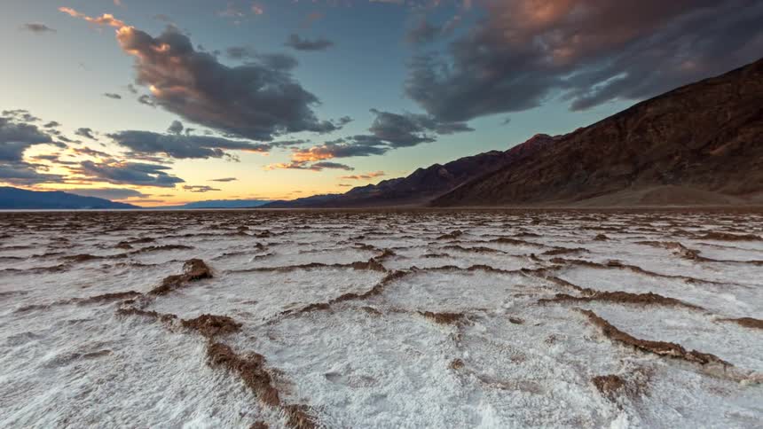 Mountains overlook snow covered rough terrain and rock formations as clouds move across the sky