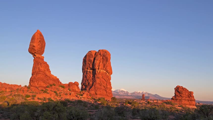 People walk on a road as sunlight shines on red rock formations in the desert with mountains in the distance