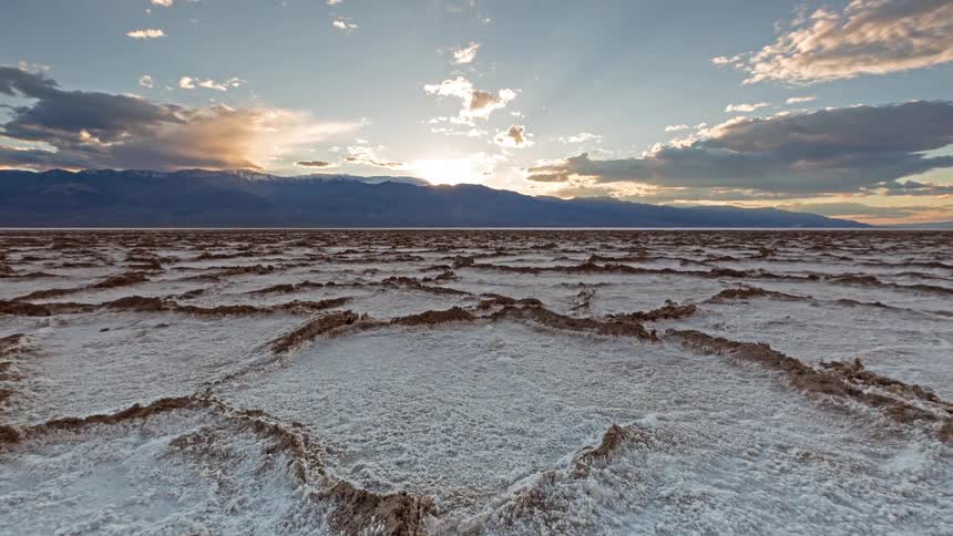 Snow covered rough terrain and rock formations as the sun shines brightly over mountains in the distance as clouds move overhead