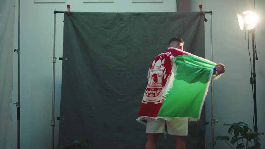 A bodybuilder poses in a studio with a middle eastern flag draped over his shoulders during a photoshoot