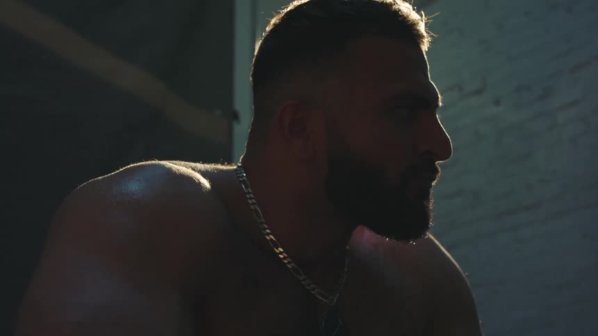 A shirtless bodybuilder sits inside a photo studio wearing a gold necklace as bright light shines on him