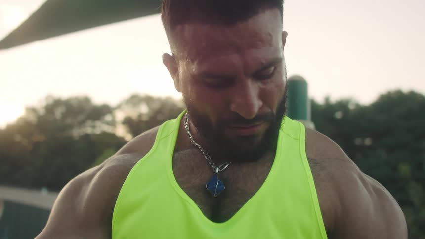 A male bodybuilder flexes his muscles outdoors at midday wearing a green singlet in the bright sun