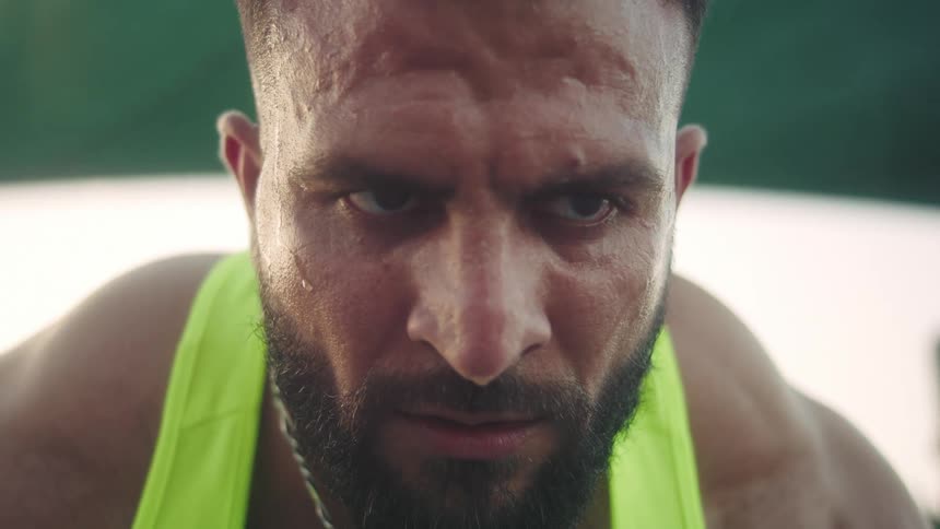 A close view of a muscular man with a beard and sweat on his skin as he looks down while wearing a neon yellow tank top and a silver chain with a blue stone