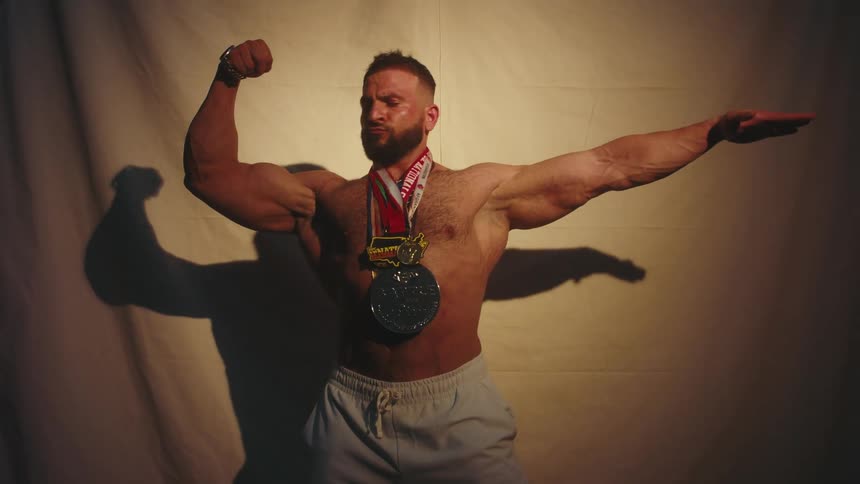 A muscular man with a beard wearing several large medals around his neck flexes his biceps and strikes bodybuilding poses in front of a plain fabric backdrop under a spotlight