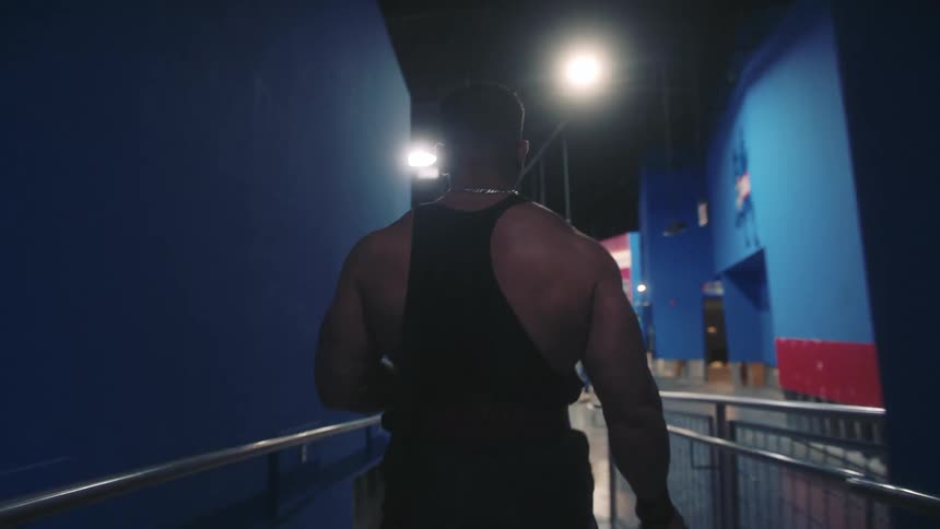 A view from the back shows a muscular man wearing a black tank top and a weight belt as he walks through a gym with blue walls toward a bright area with workout equipment