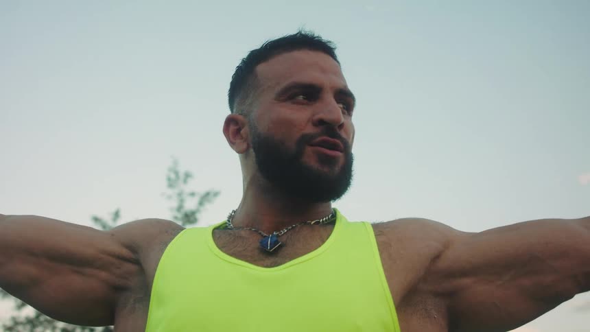 A muscular man with a beard stands outdoors under a clear sky wearing a bright yellow tanktop and earbuds