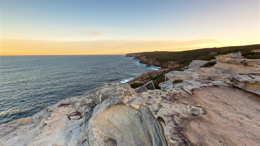 A view of a rocky coastline shows a high cliff over the ocean where the sky changes from blue and gold to purple as the sun sets and a full moon rises above the horizon