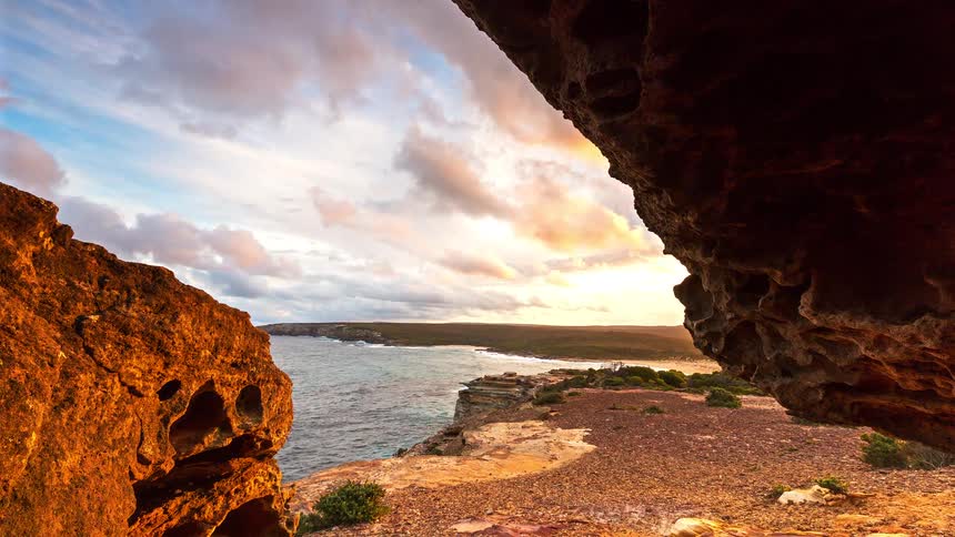 A view from inside a cave shows a rocky coastline and a large body of water under a cloudy sky as the sun begins to set