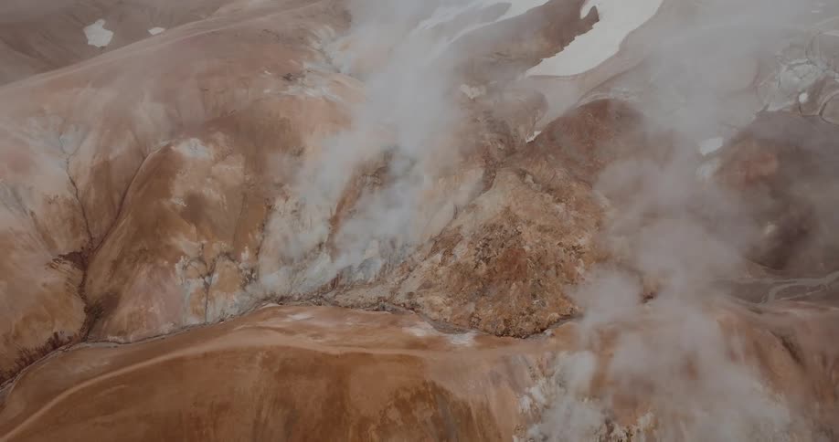 A high angle view shows steam rising from geothermal vents on brown rocky mountains with patches of white snow under a gray sky
