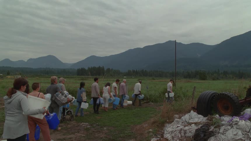 Under a heavy overcast sky a diverse group of people stand in a patient line across a grassy rural field to fill plastic jugs with water from a single ground source framed by a vast and somber mountain range