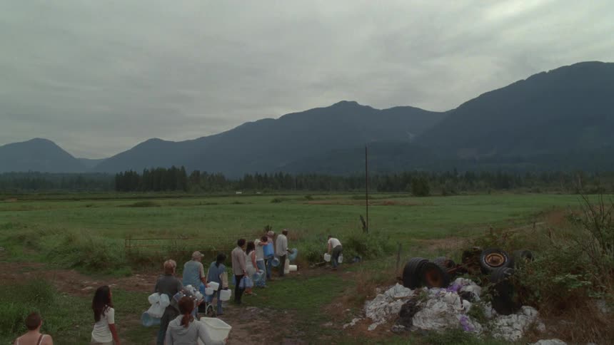 People wait in line to refill water containers from a water source in a field bordering fog covered mountains