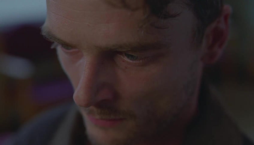 A man is shown in a tight closeup looking downward with a somber or pensive expressionas the shot continues he remains still with a furrowed brow his eyes fixed on something out of frame while his face is dimly lit