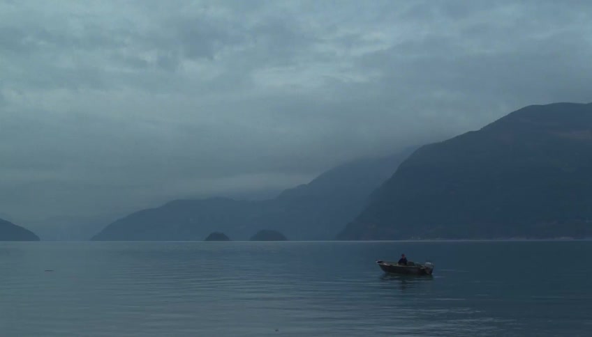 A lone person sits in a small motorized boat drifting across a vast glassy body of water under a heavy overcast skythe quiet bluetoned landscape is framed by the dark layered silhouettes of massive mountains receding into the misty horizon