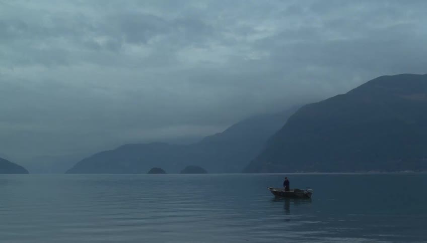 Lone boat moving across calm waters with mountains in the background under an overcast sky