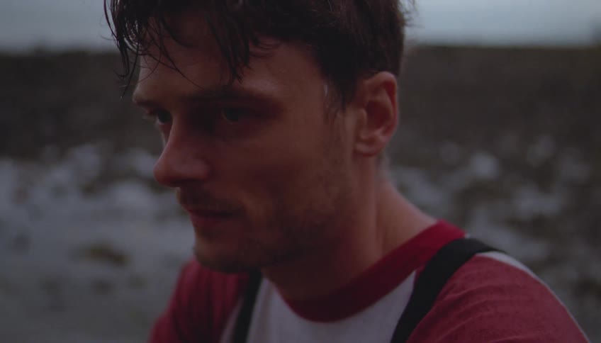 A man with wet hair and a somber expression looks downward while standing in a bleak muddy environmenthe wears a red and white shirt with black straps as he slowly lifts his gaze toward the horizon under an overcast sky