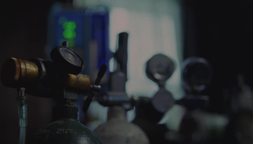 Several oxygen cylinders equipped with pressure regulators stand in the foreground while a blue digital monitor glows softly in the blurred background near a window