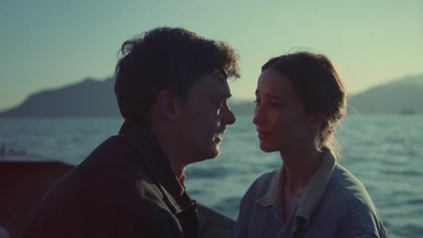 A man and a woman sit closely together on a boat sharing an intimate moment as the sun begins to set over the water and distant mountainsthe man speaks softly to the woman while they gaze at each other eventually leaning in to rest his forehead agai