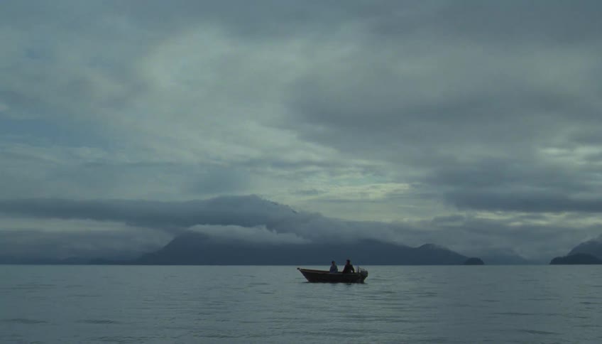 Two people float in a small boat on calm water with misty mountains and a cloudy sky