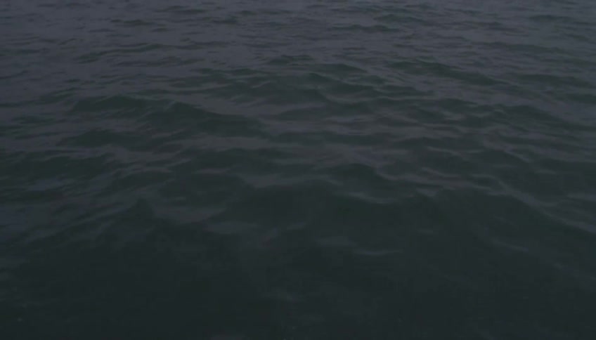 The video shows a closeup highangle view of dark choppy ocean water moving under a dim overcast sky gentle waves ripple across the surface creating a moody and rhythmic texture across the frame