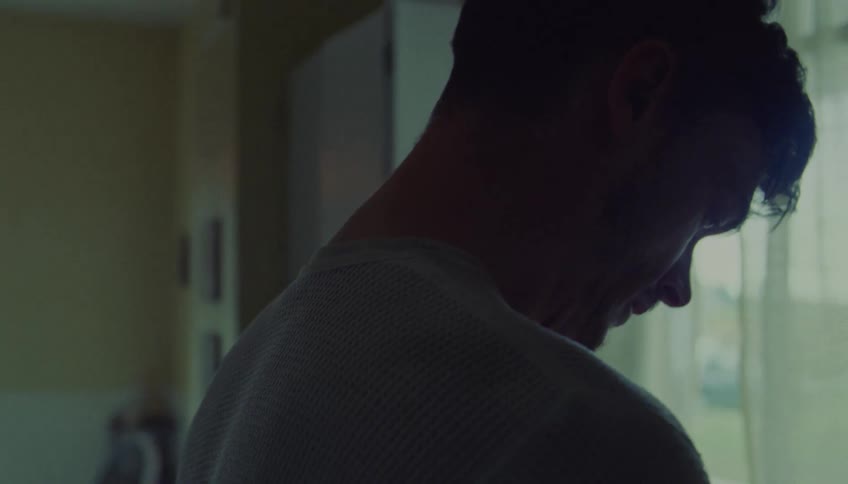 A man stands indoors with his head bowed and a slight smile on his face seen from a side profile the scene is dimly lit with soft light coming from a nearby window in the background