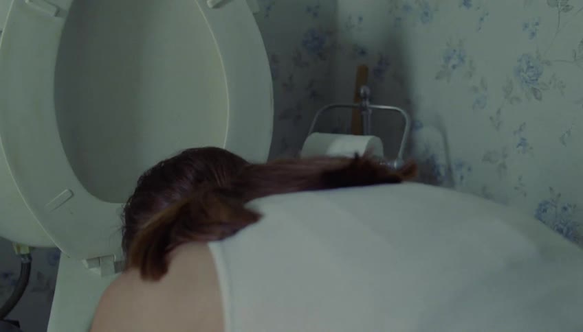A woman in a white tank top hunches over a toilet and vomits
