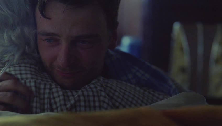 A young man with wet hair and tearful eyes intensely hugs an elderly person with gray hair while lying in bed showing a profound moment of grief and comfort as he rests his chin on their shoulder and closes his eyes in a dark room