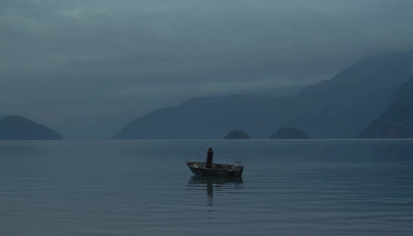Man standing in a small boat pulling a fishing net with mountains along the coast