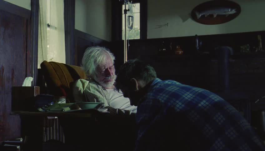 An elderly man with white hair and a nasal cannula rests in a chair while a younger man in a plaid shirt leans in and gives him a gentle hug in a dimly lit room with a mounted fish on the wall