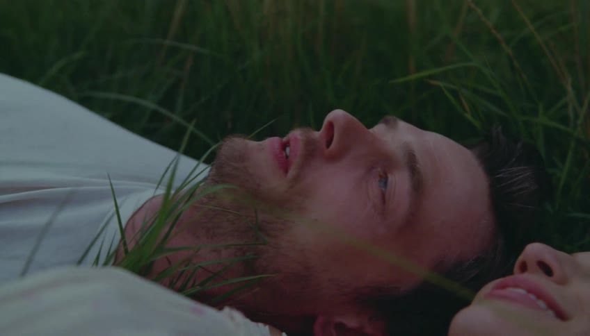 A couple lies together in tall grass looking up and smiling