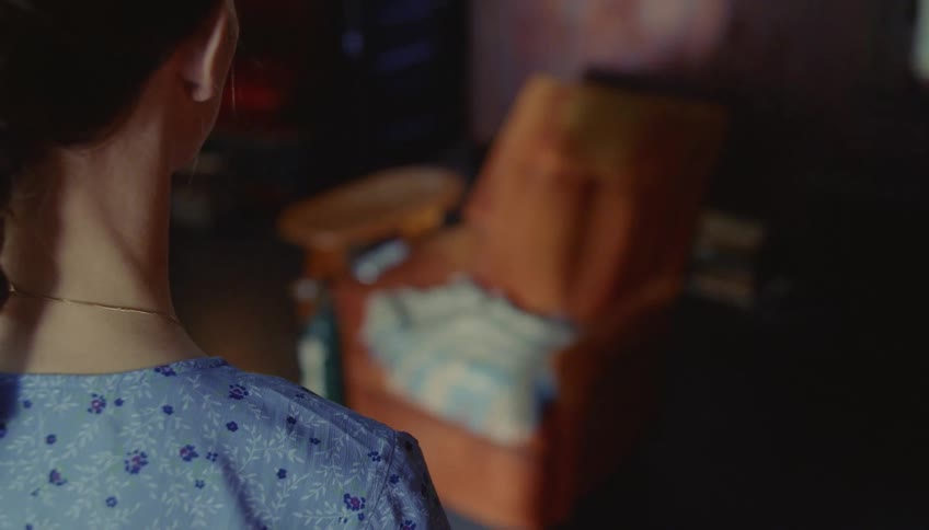 A woman in a braided ponytail walks into a dimly lit room pauses to look at an empty orange recliner with an oxygen tank beside it and then walks away
