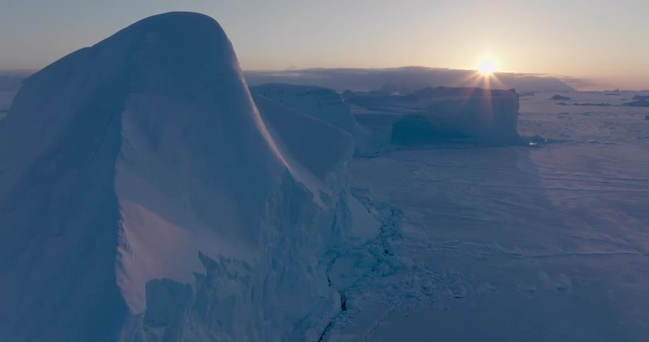 An aerial camera glides over a massive white iceberg during a polar sunrise as sunlight glints off the frozen ocean and illuminates the vast snowy landscape of the arctic circle