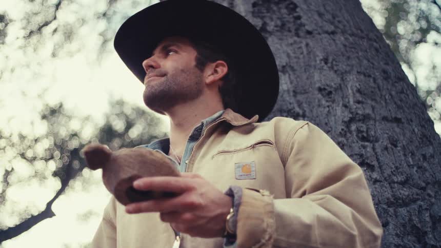 A man wearing a black hat and tan jacket leans against a large tree and smiles while holding a small wooden carving