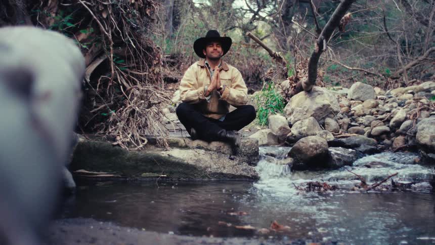 Man crouches by a stream in a wooded area with rocks and branches around