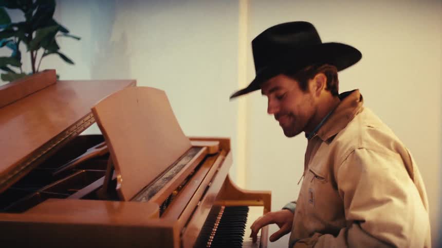 Man wearing a hat plays a piano in an indoor setting with warm lighting