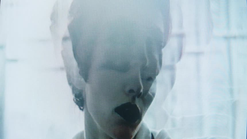 A woman with dark lipstick and short hair moves her head in a stylized dreamlike manner behind a sheer hazy veil the footage has a distorted lofi aesthetic with visible scan lines and lens flares that create a ghostly atmosphere
