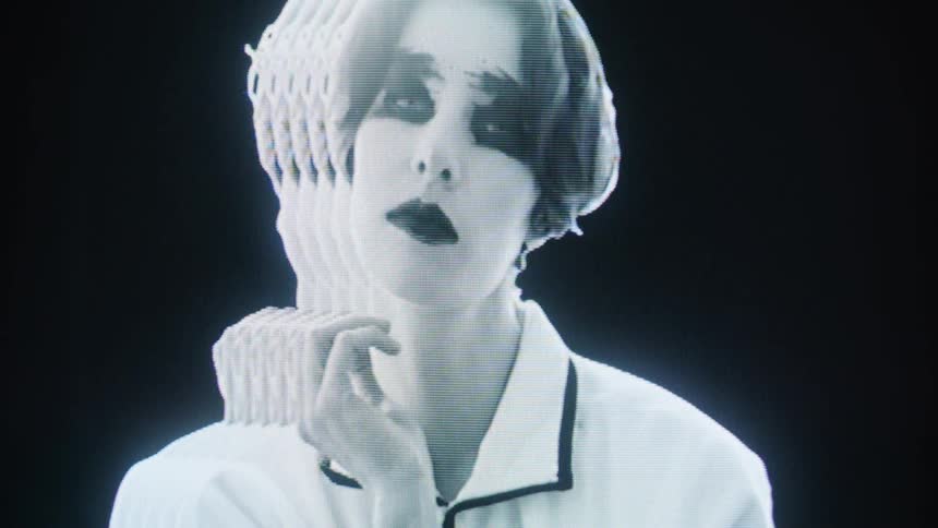 A person with dramatic dark eye makeup and dark lipstick poses in front of a glitchy crtstyle screen an echo effect creates multiple ghostly silhouettes that trail behind their head and hand movements