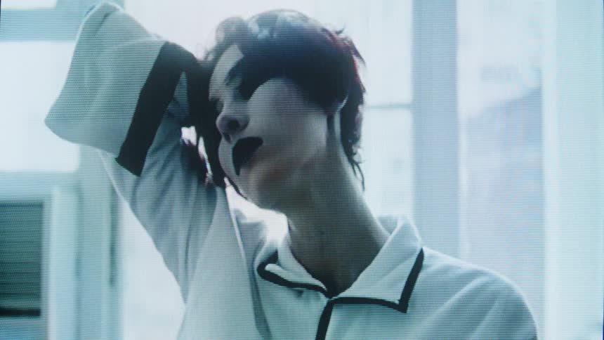 Woman with dark makeup touches her face with raised arms in an indoor space lit by a window