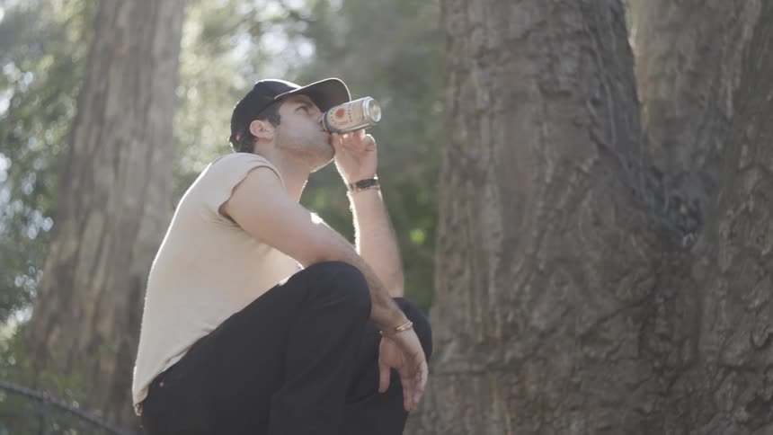 A man wearing a black baseball hat and a white shirt crouches in a wooded area and takes a drink from a silver can while sunlight filters through the trees