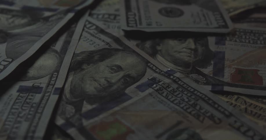 A close view shows several one hundred dollar bills featuring the portrait of benjamin franklin scattered together in a pile under dim light