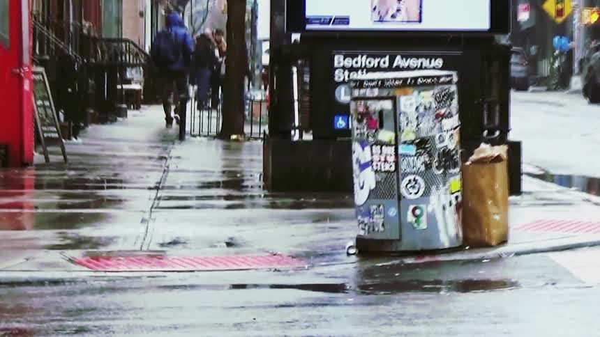 A close view of a yellow taxi driving past a wet city sidewalk near a subway entrance on a rainy day